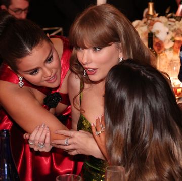 selena gomez hugs taylor swift who is sitting at a table and talking, gomez wears a red dress and swift wears a green sequin dress