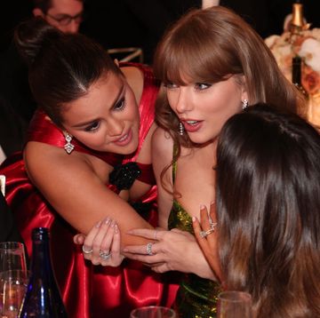 selena gomez hugs taylor swift who is sitting at a table and talking, gomez wears a red dress and swift wears a green sequin dress
