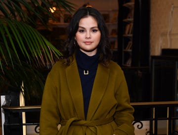 selena gomez looks directly at the camera, she wears a black turtle neck with a gold detail and an olive green coat, behind her are bookshelves and a tropical plant