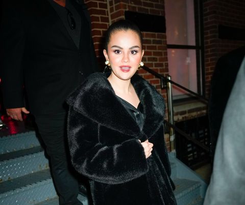 selena gomez looks at the camera with her mouth slightly open, she wears a long black fur coat, large sparkling earrings, and makeup with her hair pulled back