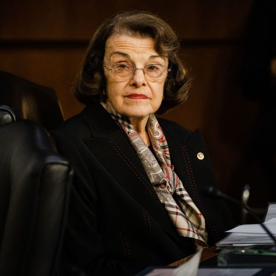 senator dianne feinstein sits at a desk in a senate chamber and looks to the left, she wears a black jacket, colorful scarf and glasses