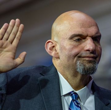 john fetterman, wearing a gray suit and tie, looks off camera and hojlds his right hand in the air