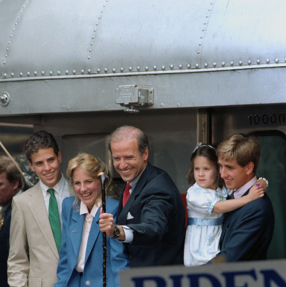 joe biden stands outside and smiles with his family including two teenage boys, a woman, and a young girl held by one of the boys