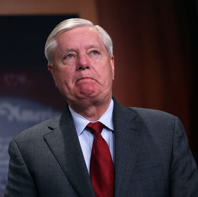 Lindsey Graham lindsey graham wearing a gray suit and red tie, frowning and looking off camera, with an american flag behind him
