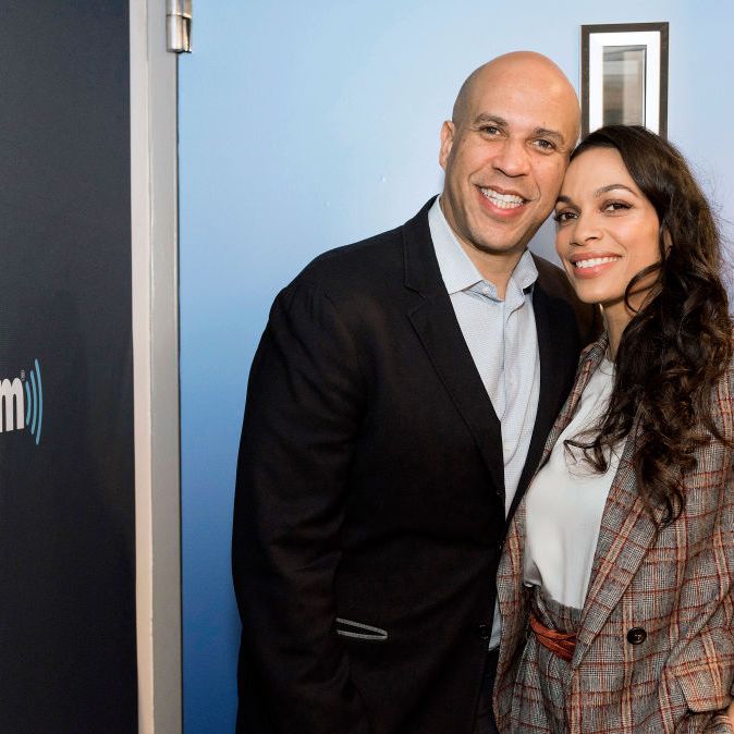 cory booker and rosario dawson embrace and smile at the camera while standing in front of a blue wall