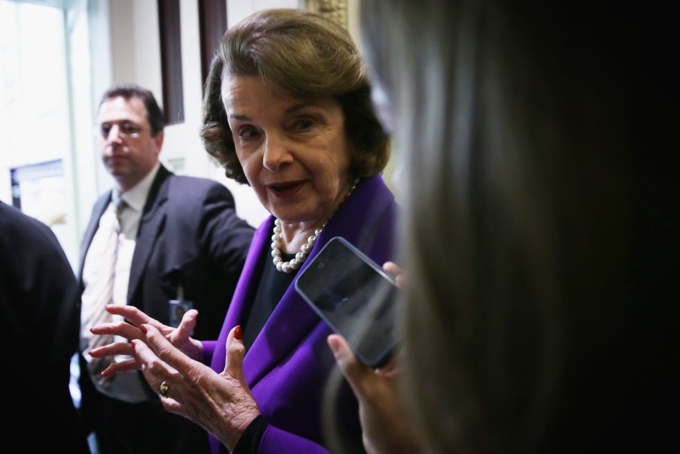 dianne feinstein, wearing a purple outfit, speaking to a reporter who uses her smartphone as a tape recorder