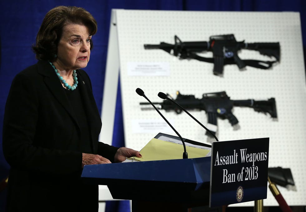 dianne feinstein speaking into a microphone at a podium, with assault weapons mounted on a display behind her