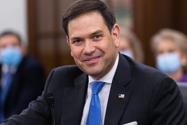 marco rubio smiles at the camera, he wears a navy suit jacket, white collared shirt and blue patterned tie, an american flag pin is on his jacket lapel