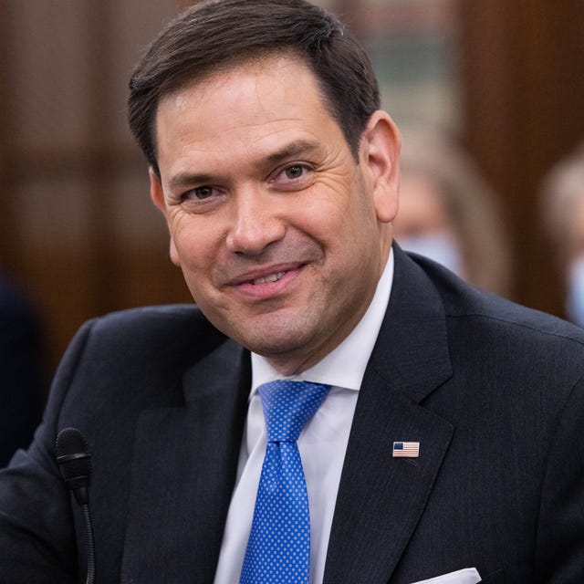 Marco Rubio in April 2021 marco rubio smiles at the camera, he wears a navy suit jacket, white collared shirt and blue patterned tie, an american flag pin is on his jacket lapel
