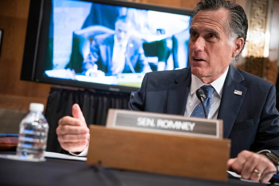 mitt romney, wearing a black suit and blue tie, sits at a table with a nameplate and bottle of water in front of him, and a television behind him