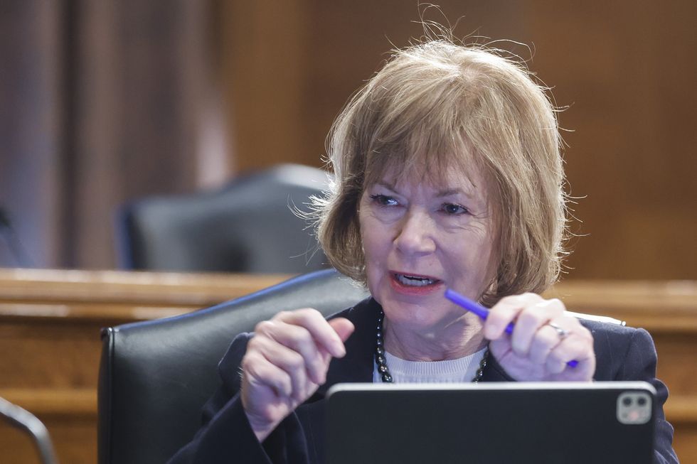 tina smith, wearing a business jacket and white shirt, speaks while sitting in a black chair in front of an open computer, holding a pen in her left hand