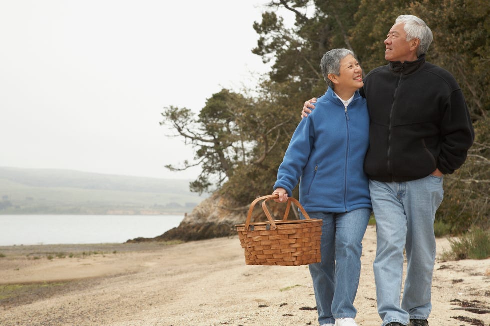 senior asian couple walking on beach with picnic basket