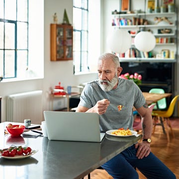 senior man sitting in kitchen eating breakfast and looking at laptop