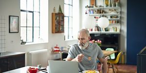 senior man sitting in kitchen eating breakfast and looking at laptop