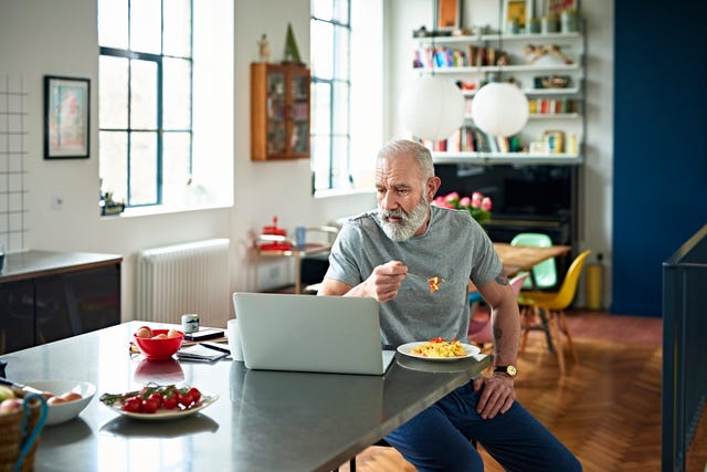 senior man sitting in kitchen eating breakfast and looking at laptop