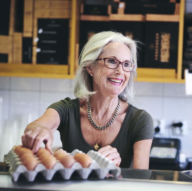 senior saleswoman smiling while picking brown egg from carton in deli