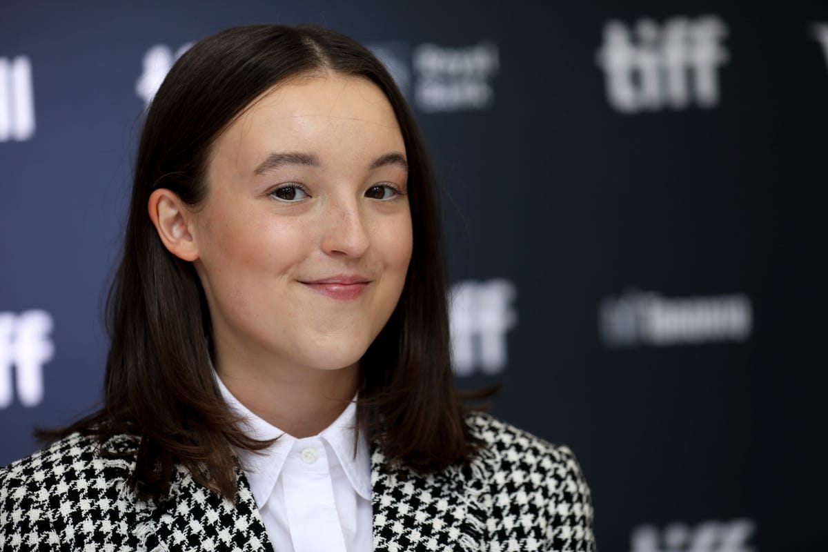 bella ramsey, smiling, looking directly into the camera, wearing a black and white patterned jacket and white shirt