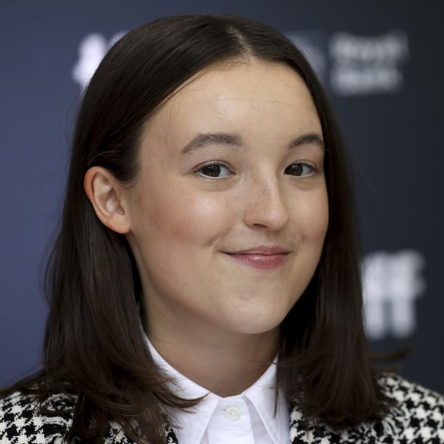 bella ramsey, smiling, looking directly into the camera, wearing a black and white patterned jacket and white shirt