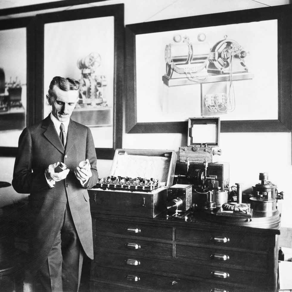 Nikola Tesla mira un dispositivo que sostiene en sus manos, está de pie con traje en una habitación con dibujos enmarcados en la pared, hay un gabinete con mucha maquinaria encima