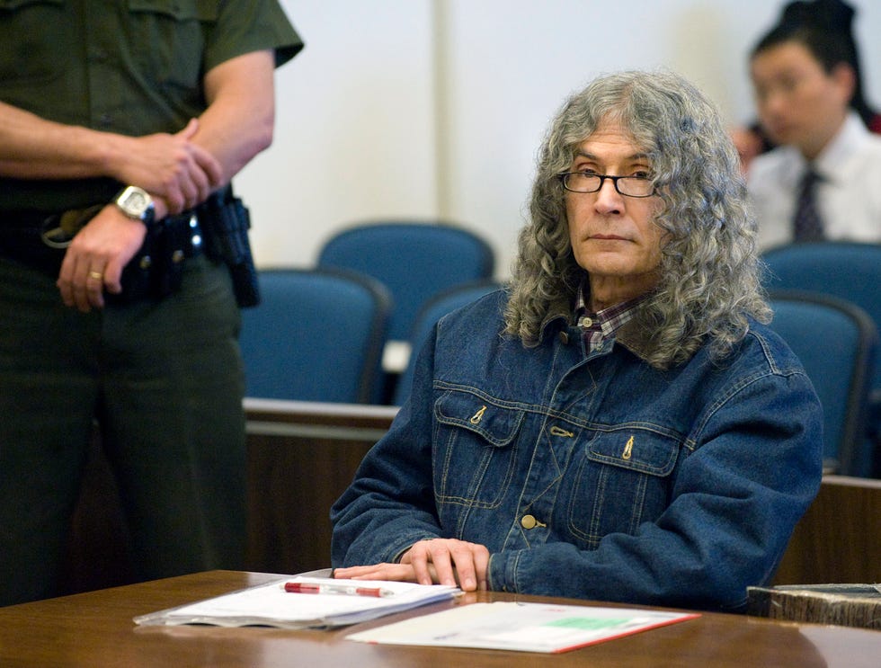 rodney alcala sits with his hands resting on a wooden table in front of him, he wears a collared plaid shirt, a denim jacket, and glasses