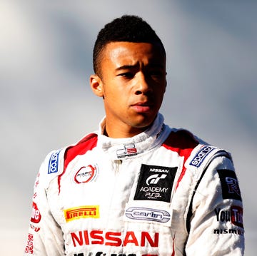 jann mardenborough stands and looks to the right of the camera with a slight frown on his face, he wears a white driver jacket with red accents and several brand logos