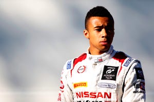 jann mardenborough stands and looks to the right of the camera with a slight frown on his face, he wears a white driver jacket with red accents and several brand logos