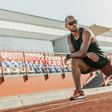 serious male athlete in sunglasses standing on the running track at the stadium while stretching his legs