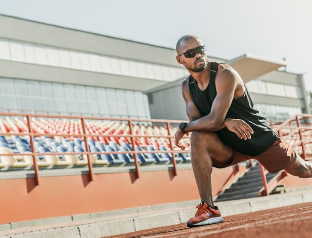 serious male athlete in sunglasses standing on the running track at the stadium while stretching his legs