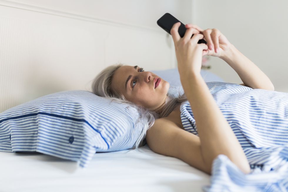 Serious young woman lying in bed checking cell phone