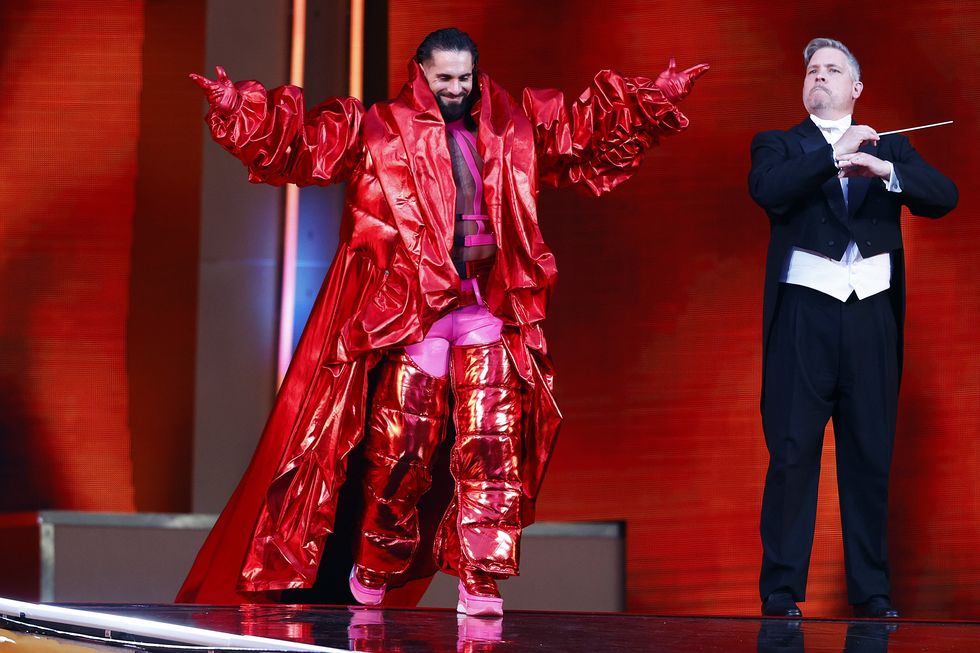 Seth Rollins sale al escenario con los brazos en alto mientras viste una chaqueta y pantalones largos de color rojo metalizado.