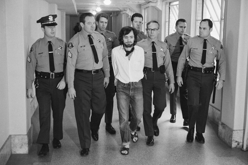 seven police officers in uniform escort charles manson as the group walks down a hallway, manson is handcuffed behind the back and wears a white shirt, jeans, and a bandana
