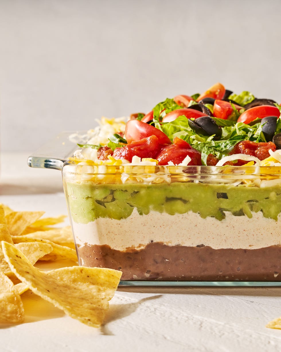 layered dip with refried beans, sour cream, guacamole, cheese, tomatoes black olives, and lettuce in a clear dish