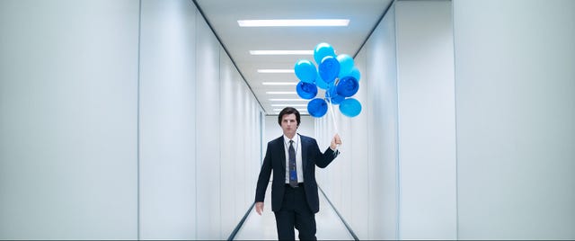 man in a suit holding blue balloons while walking in a modern hallway