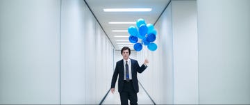 man in a suit holding blue balloons while walking in a modern hallway