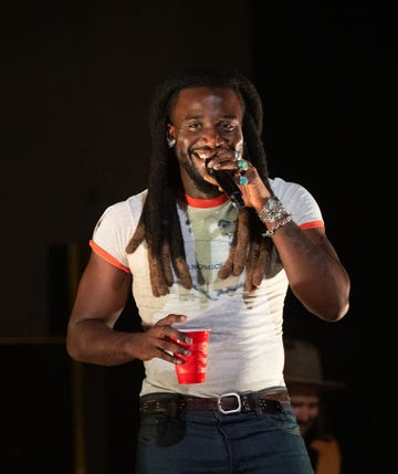 shaboozey smiles as he holds a wireless microphone to his mouth, he holds a red plastic cup in his other hand and wears a graphic tshirt and jeans