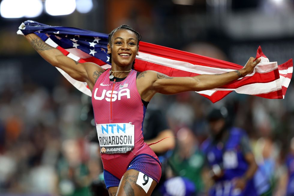 shacarri richardson smiles and looks past the camera while holding an american flag behind her back with outstretched arms, she wears a pink and blue uniform with a bib on her stomach