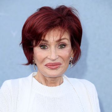 a woman with short red hair smiles at the camera, she wears a white sweater set and dangling diamond earrings