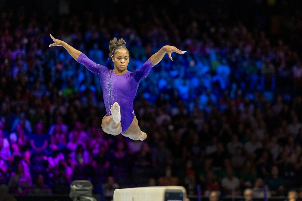 shilese jones jumps while competing on the balance beam, her arms are extended at her sides and her legs are extended in a forward split, she wears a purple leotard and looks down, a crowd is in the background