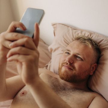 shirtless obese man taking selfie through smart phone while lying on bed at home