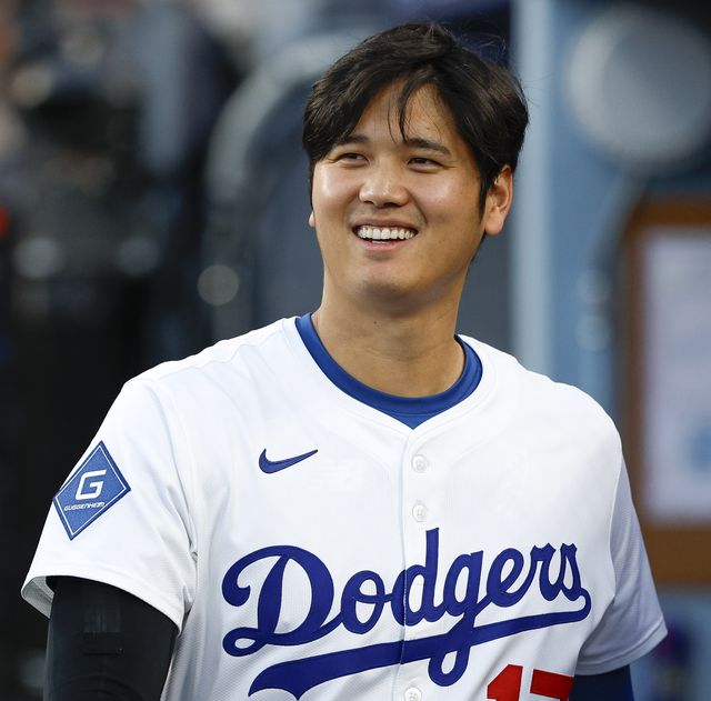 a man with dark hair smiles and looks left, he wears a los angeles dodgers baseball uniform that's white and blue