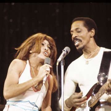 a woman and a man stand close together while performing on a stage, they wear white outfits and sing into microphones, he plays electric guitar
