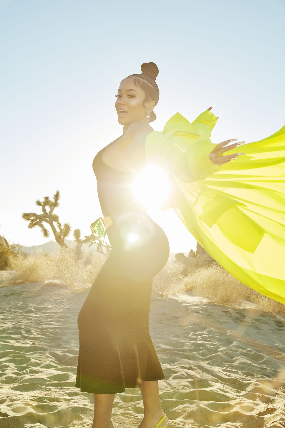 singer saweetie, wearing a dress, coat, and fanny pack, with hands up behind her with coat flying in the wind, in front of a sky and desert backdrop