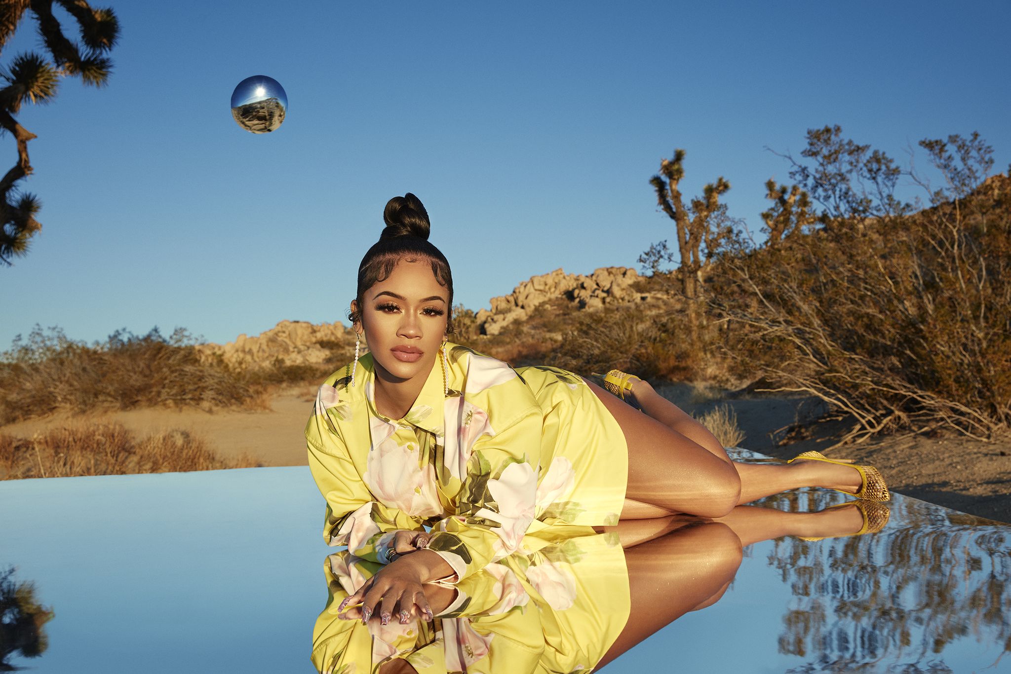 singer saweetie lying down on top of a large reflective mirror, wearing a yellow dress and heels, in front of a desert and sky background with a reflective silver globe in the sky