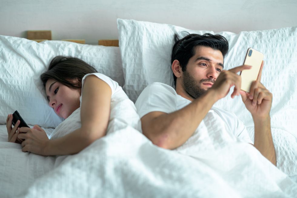 shot of a young couple using their cellphones in bed at evening back to back