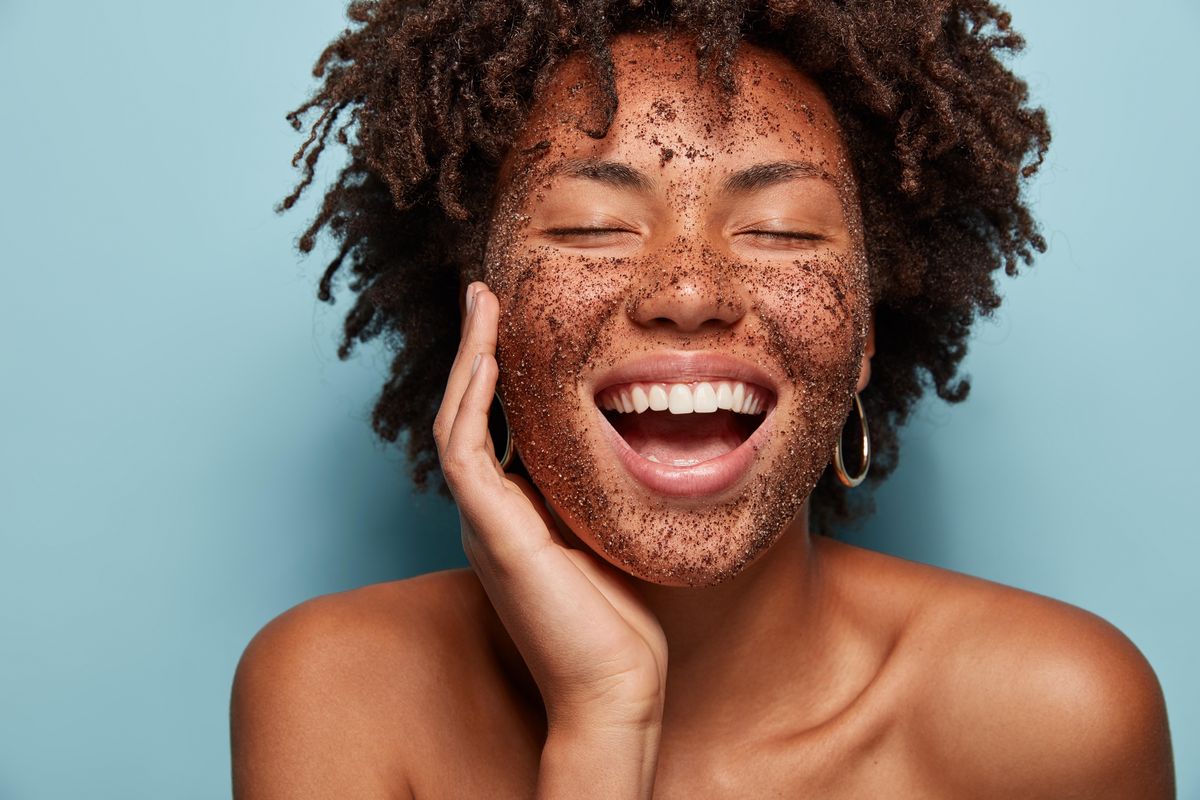satisfied african american woman has pore cleaning with scrub mask, laughs happily and shows white teeth, has curly hair, healthy skin, isolated over blue studio wall, uses peelig cosmetic product shutterstock id 1314987779 brand   issue   ordered by