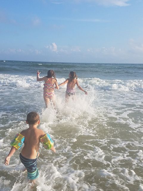 three siblings at beach running into low surf with blue sky in background