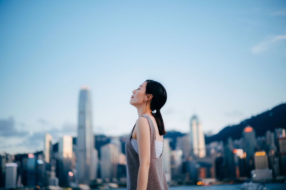 side profile of young asian woman taking a deep breath with her eyes closed, enjoying the fresh air against urban cityscape in downtown district in city