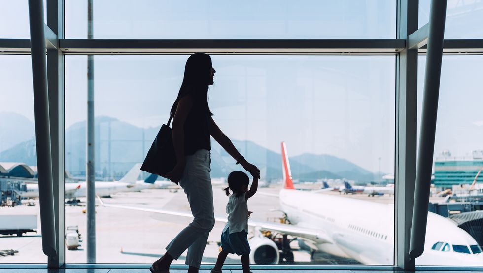 silhouette of joyful young asian mother holding hands of cute little daughter looking at airplane through window at the airport while waiting for departure