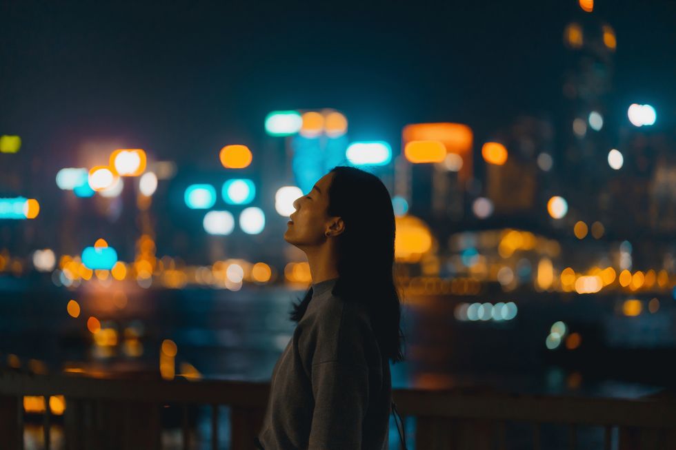 silhouette of side profile of a young asian woman standing against illuminated and colourful bokeh lights background of urban cityscape at night