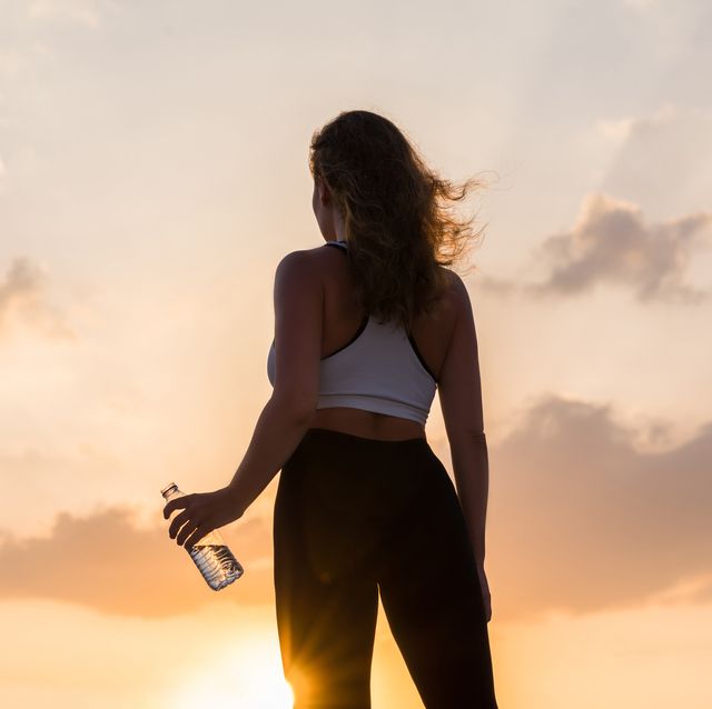 silhouette of young woman in the sport clothes with bottle of clear mineral water on the sunset sky backgrounds the concept of healthy lifestyle